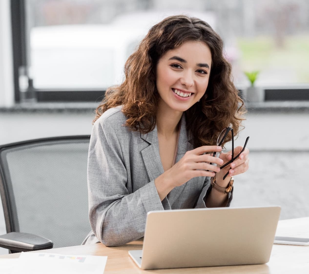 Girl sitting infront of laptop smiling while holding a pair of glasses